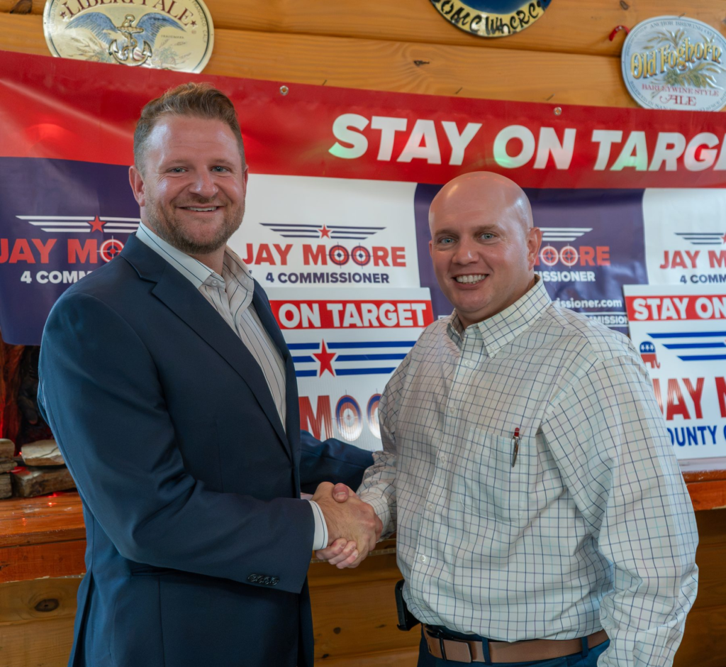 Jay Moore, bald in a light windowpane-check shirt, shaking hands with a neighbor in a navy blazer in front of Stay on Target banners
