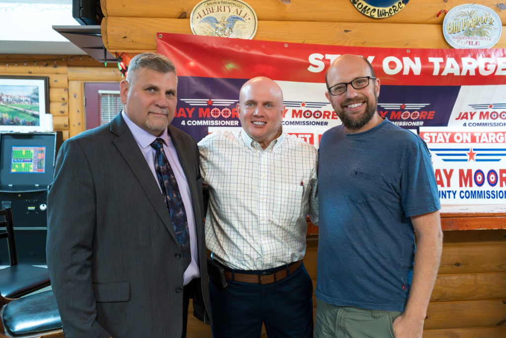 Jay Moore, bald in a light windowpane-check shirt, standing with two neighbors in front of Stay on Target banners