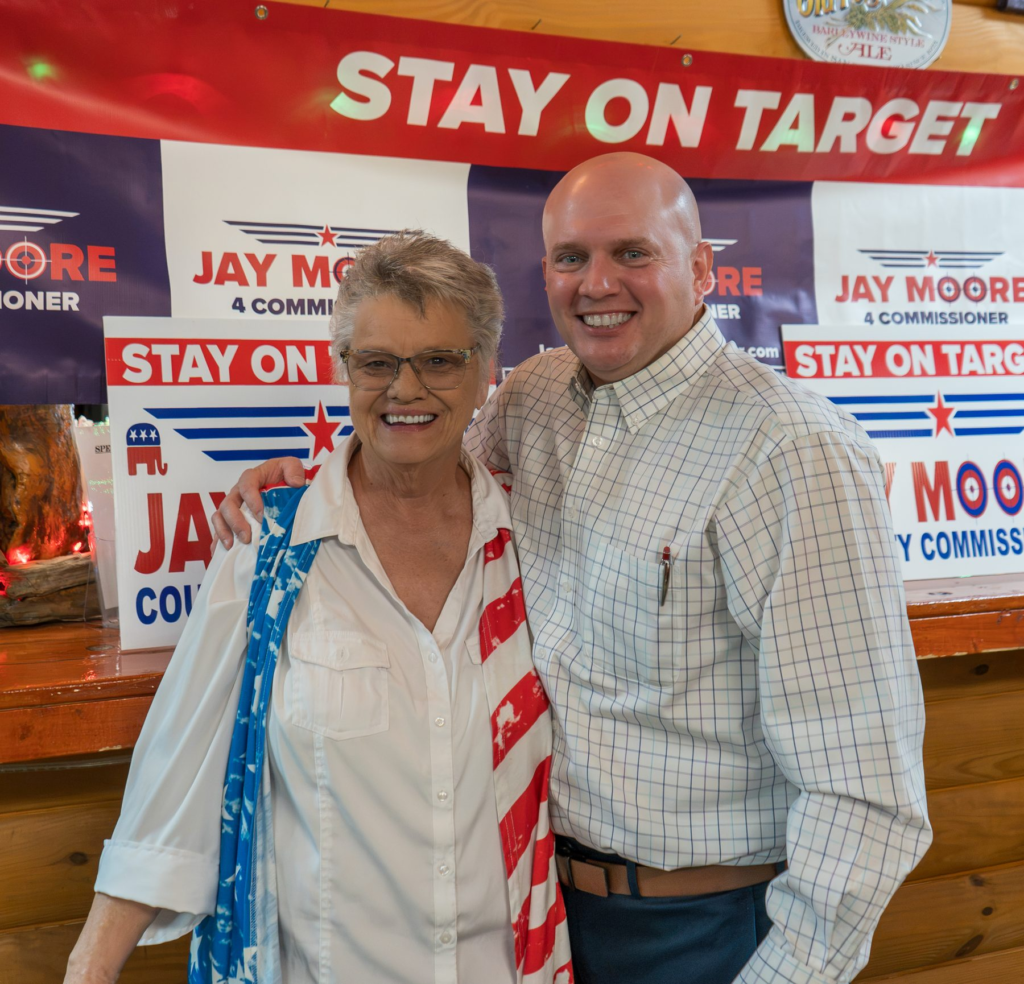 Jay Moore, bald in a light windowpane-check shirt, with a neighbor in an American flag scarf at a Stay on Target campaign event
