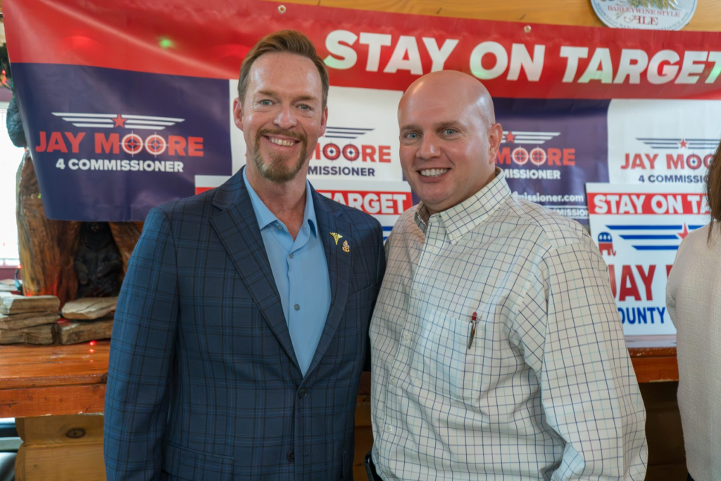 Jay Moore, bald in a light windowpane-check shirt, standing with a neighbor in a navy blazer in front of Jay Moore 4 Commissioner signs