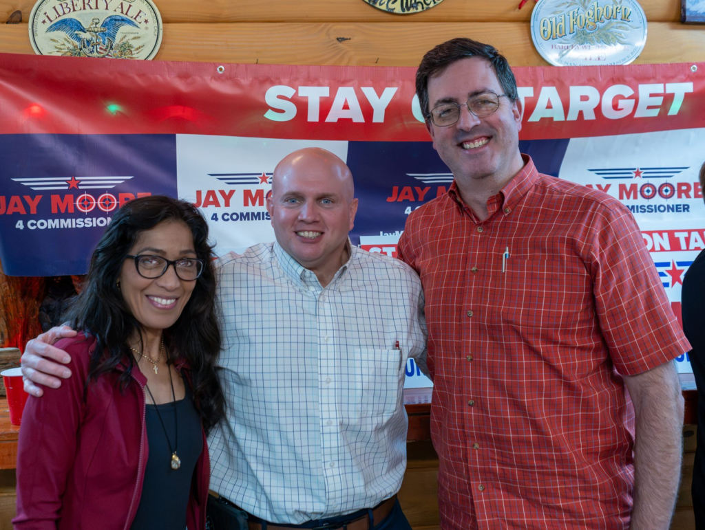 Jay Moore, bald in a light windowpane-check shirt, with two neighbors at a local campaign gathering