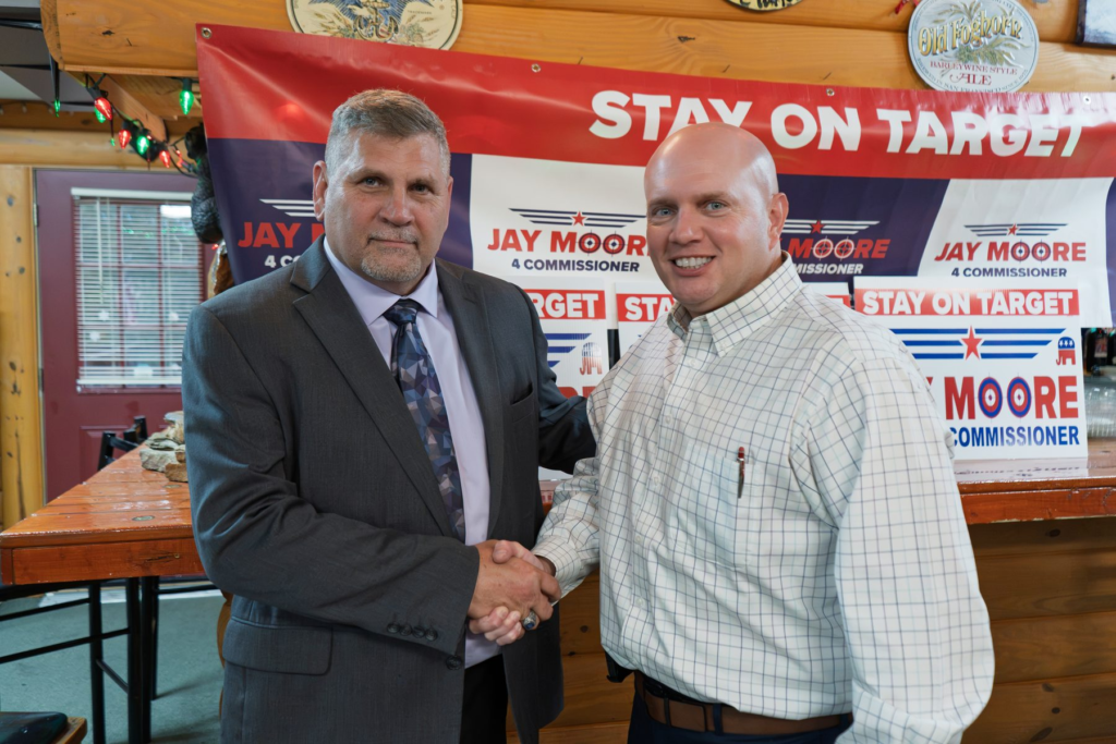 Jay Moore, bald in a light windowpane-check shirt with a pen in his pocket, shaking hands with a neighbor in a suit