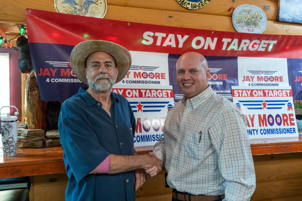 Jay Moore, bald in a light windowpane-check shirt, shaking hands with a neighbor in a straw hat in front of Stay on Target signs