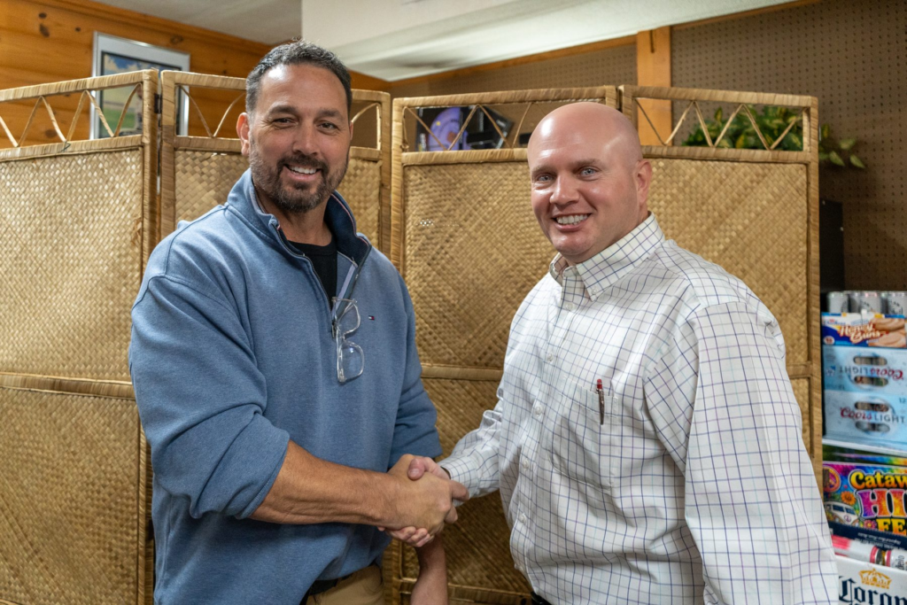 Jay Moore, bald in a light windowpane-check shirt, shaking hands with a neighbor in a blue quarter-zip at a local business