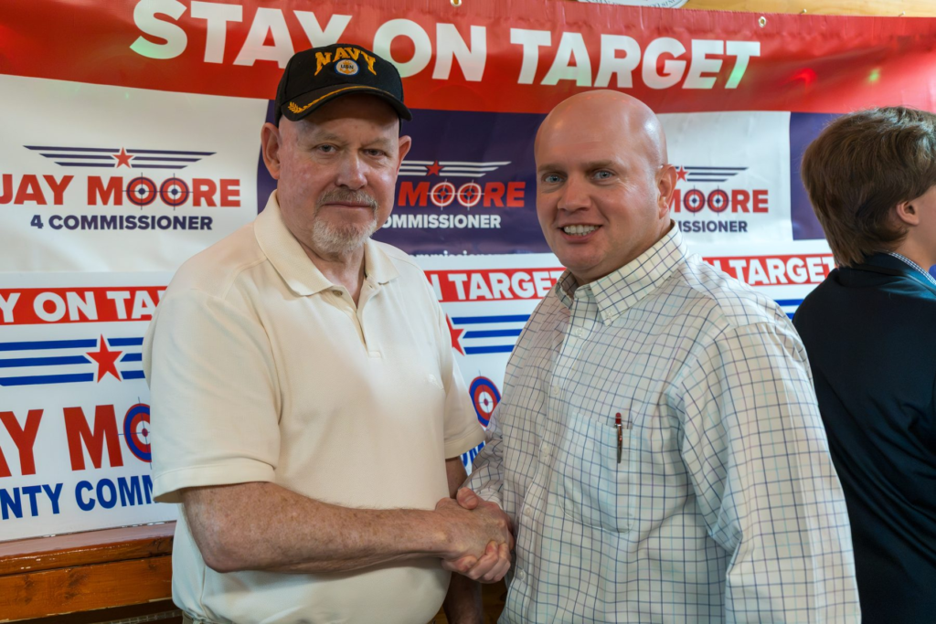 Jay Moore, bald in a light windowpane-check shirt, shaking hands with a neighbor in a navy cap